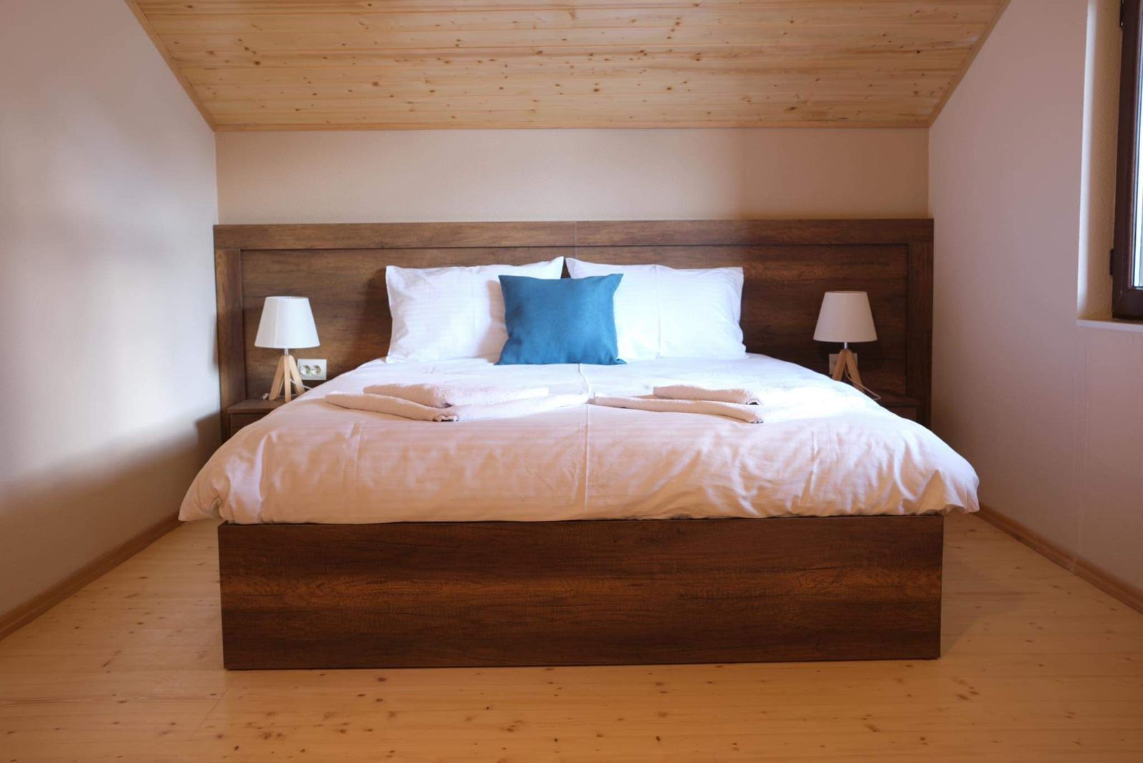 Un lit bien fait, avec du linge de lit blanc et un oreiller bleu, deux lampes de chevet et une tête de lit en bois.