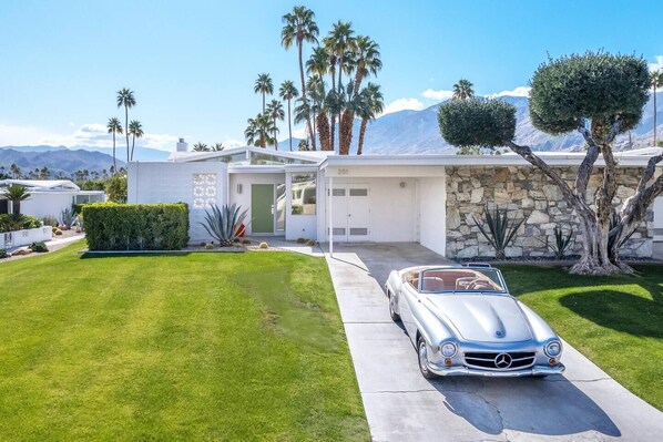 Une maison moderne avec une façade en pierre, une pelouse bien entretenue et une voiture décapotable classique garée dans l’allée.