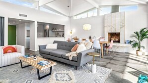Amplia sala de estar con un sofá gris, una mesa de centro y una mesa de comedor con sillas. Se observa una cocina al fondo, una chimenea y un ventanal que deja entrar luz natural.