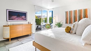 Un dormitorio moderno con una cama grande, televisor, cómoda de madera y puerta corrediza de vidrio que da a un balcón con vistas a las montañas.