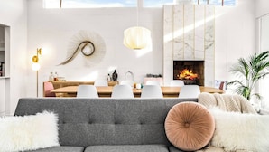 Una sala de estar moderna con un sofá gris, un comedor con mesa de madera y sillas blancas, una chimenea y un ventanal que proporciona luz natural.