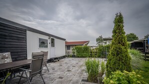 A patio area with a wooden table and chairs, a white building, and a tall coniferous tree. - Modern chalet on the Wadden Sea, near the beach (Westerland)