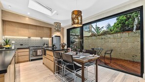 A modern kitchen with a dining area, featuring a large island, wooden cabinetry, and a view of the outdoors. - North Fremantle Cottage (North Fremantle)