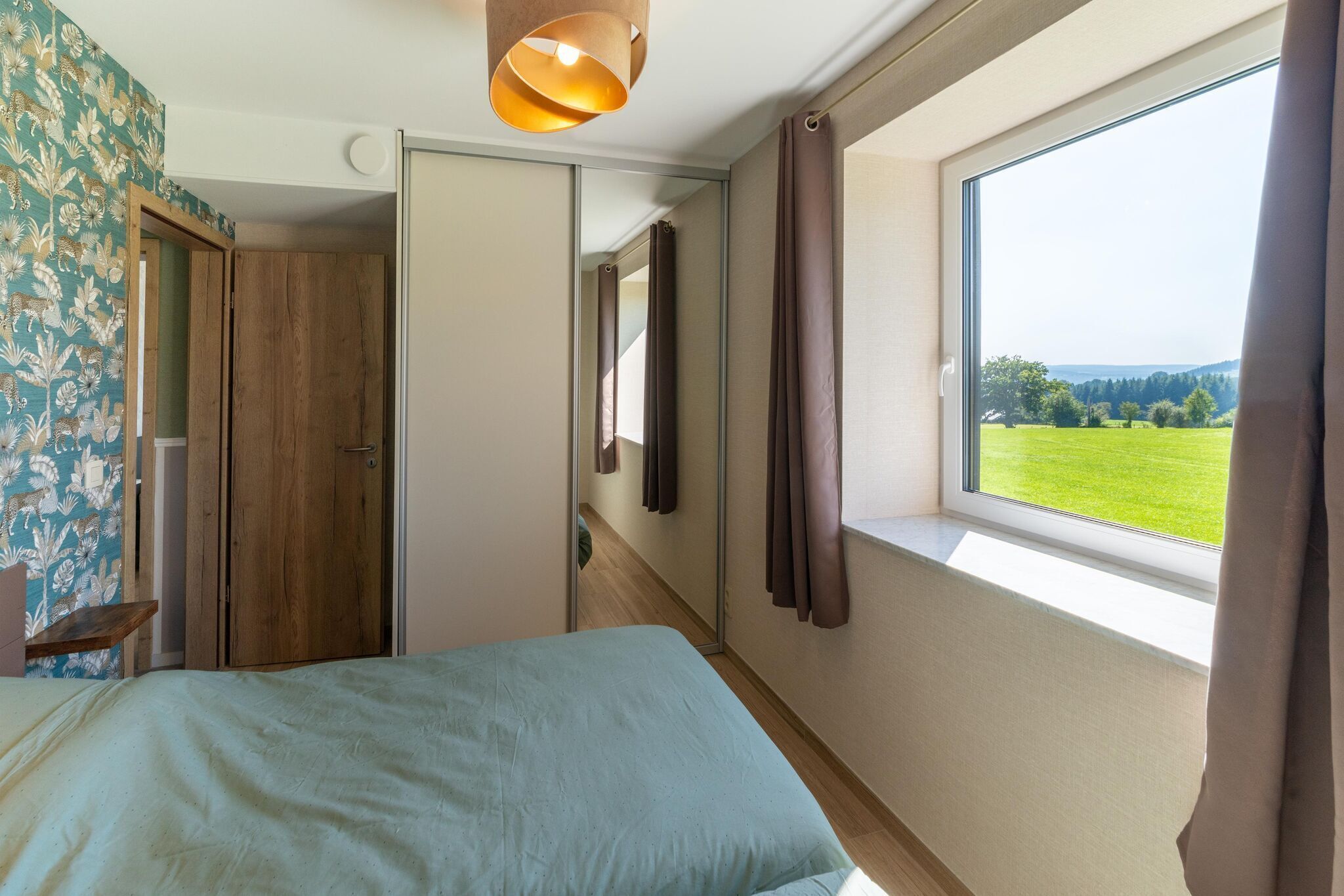 A bedroom with a bed, a window with a view of a green field, a wooden wardrobe, and a mirror.
