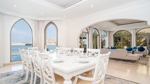 A spacious dining area with a white table and chairs, large windows with a view of the sea, and a modern living area with a sofa and cushions. - 5BR Beach & pool villa in Palm (Dubai)