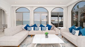 A spacious living room with a beige sofa, blue and white patterned pillows, a white coffee table, and large arched windows offering a view of the sea. - 5BR Beach & pool villa in Palm (Dubai)
