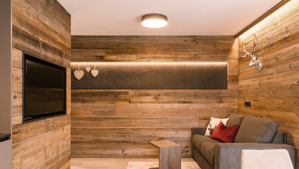 Ein modernes Wohnzimmer mit Holzwandverkleidung, einem Flachbildfernseher, einem grauen Sofa mit roten und weißen Kissen und einem kleinen Holztisch.