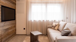 A living room with a flat-screen TV mounted on a wooden wall, a beige sofa with decorative pillows, a small wooden side table, and sheer curtains on the window.