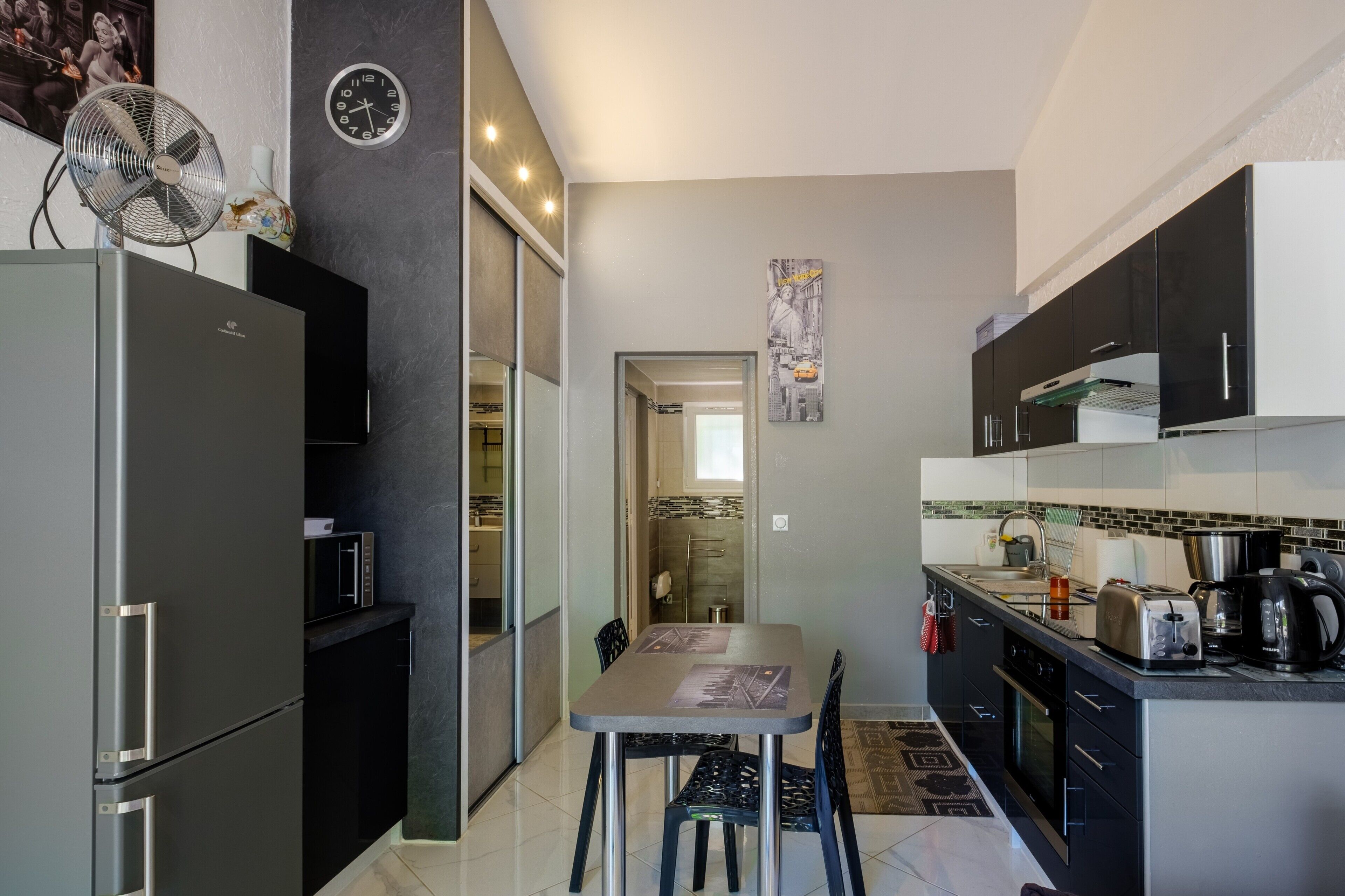 A compact kitchen with a refrigerator, microwave, and stove. There is a dining table with chairs and a fan.