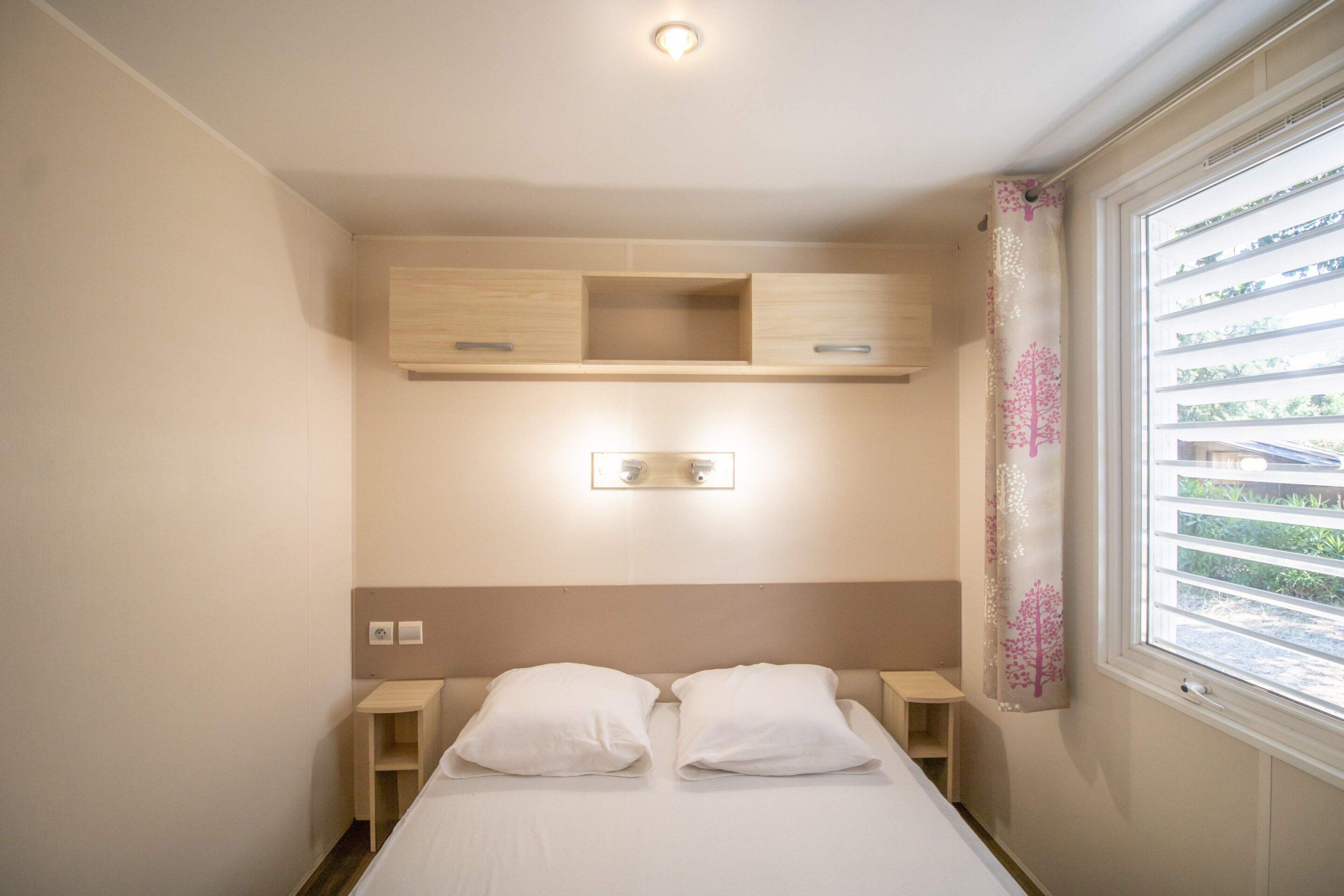 Une chambre à coucher avec un lit, des tables de chevet, une lampe fixée au mur et une fenêtre avec des stores.