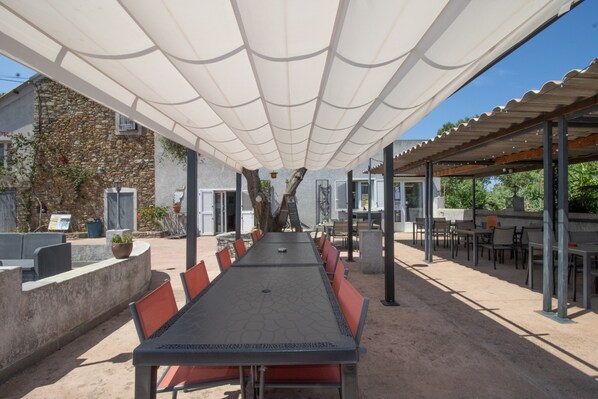 An outdoor dining area with a long table and chairs, shaded by a white canopy, and a stone building in the background.