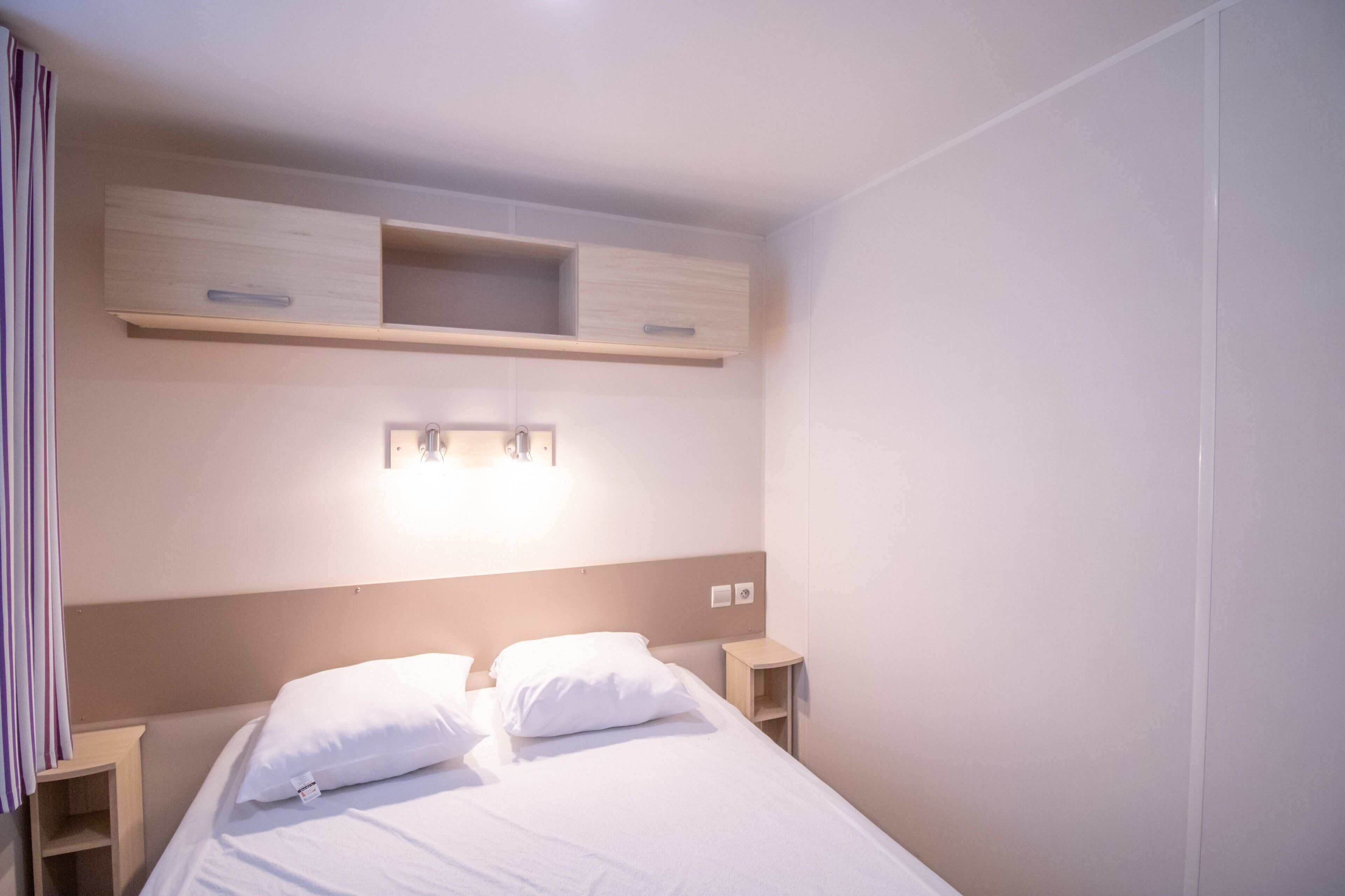 A hotel room with a bed, bedside tables, a light fixture above the bed, and a wall-mounted shelf.