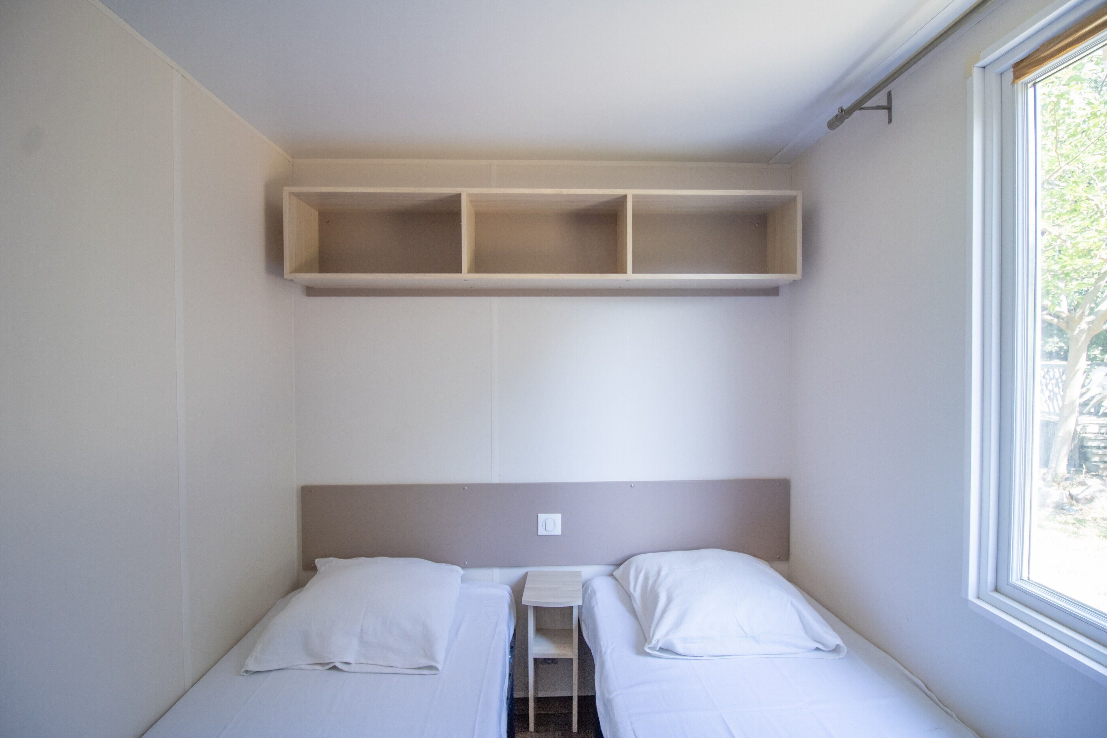 Ein kleines Zimmer mit einem Einzelbett, einem Fenster, einem eingebauten Regal über dem Bett und einer Steckdose an der Wand.
