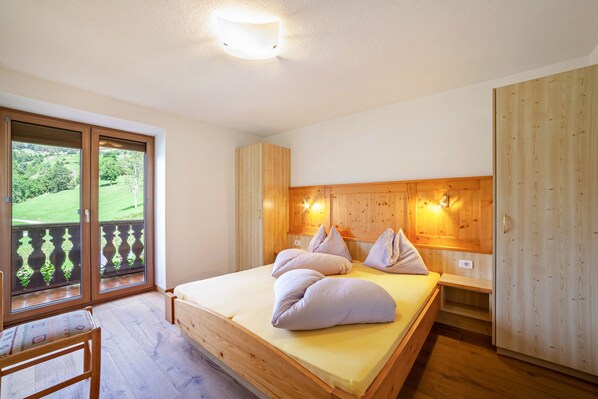 A bedroom with a wooden bed, a nightstand, a wooden wardrobe, and a balcony with a view of greenery.