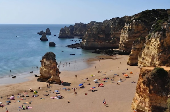 A beach with rocky cliffs, clear blue water, and people sunbathing.