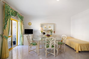 Un coin repas avec une table en verre, des chaises blanches, un téléviseur et un lit jaune.