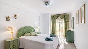 Un dormitorio con cabecera verde, cama con ropa de cama blanca, mesita de noche con lámpara, cómoda y ventana con cortinas.