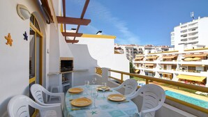 Un balcón con una mesa dispuesta para comer, sillas de plástico blanco y vistas a una piscina y edificios al fondo.