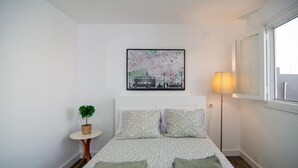 Een slaapkamer met een bed, kussens, een kaart als kunstwerk, een lamp, een klein tafeltje met een plant, en een raam.