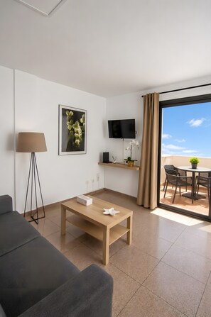 Habitación de hotel moderna con sofá, mesa de centro, televisor, televisor de pantalla plana en la pared, una mesita con planta y balcón con mobiliario exterior.