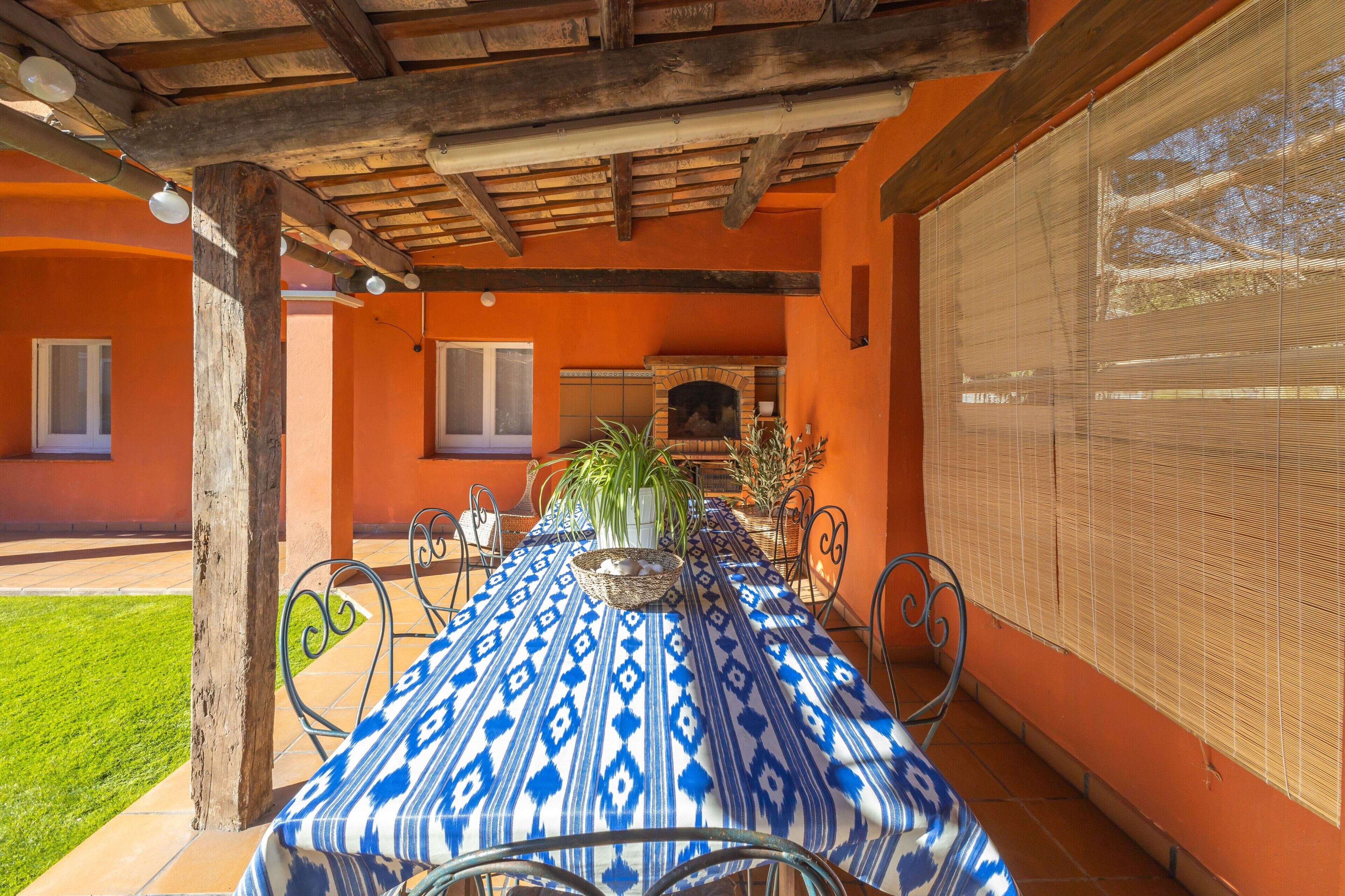 An outdoor dining area with a patterned tablecloth, metal chairs, a wooden pergola, and a brick fireplace.