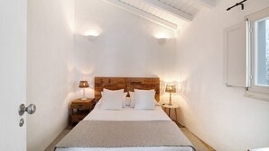 Un dormitorio con cabecera de madera, dos mesitas de noche con lámparas, una cama con ropa blanca y un ventanal con vistas.