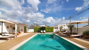 Una piscina rectangular con un muelle de madera, rodeada de sillas de descanso y sombrillas, bajo un cielo despejado.