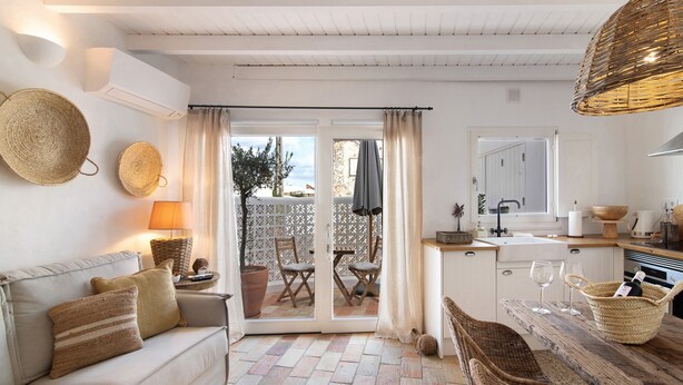 Un espacio de sala acogedor con un sofá, una mesa de comedor, sillas de mimbre, una zona de cocina y un ventanal corredizo que da a un patio exterior.
