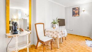 Een kamer met een grote spiegel, een koffiezetapparaat, een rieten stoel, een tafel met een bloemendekbed en een schilderij aan de muur.