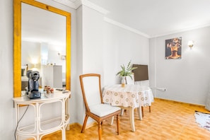 Une pièce avec un grand miroir, une machine à café, un fauteuil en osier, une table recouverte d’une nappe à motifs floraux et un tableau accroché au mur.