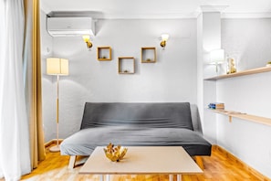 Un salon moderne avec un canapé gris, une table basse en bois et un bol décoratif. On y trouve des lampes fixées au mur et une étagère remplie de livres.