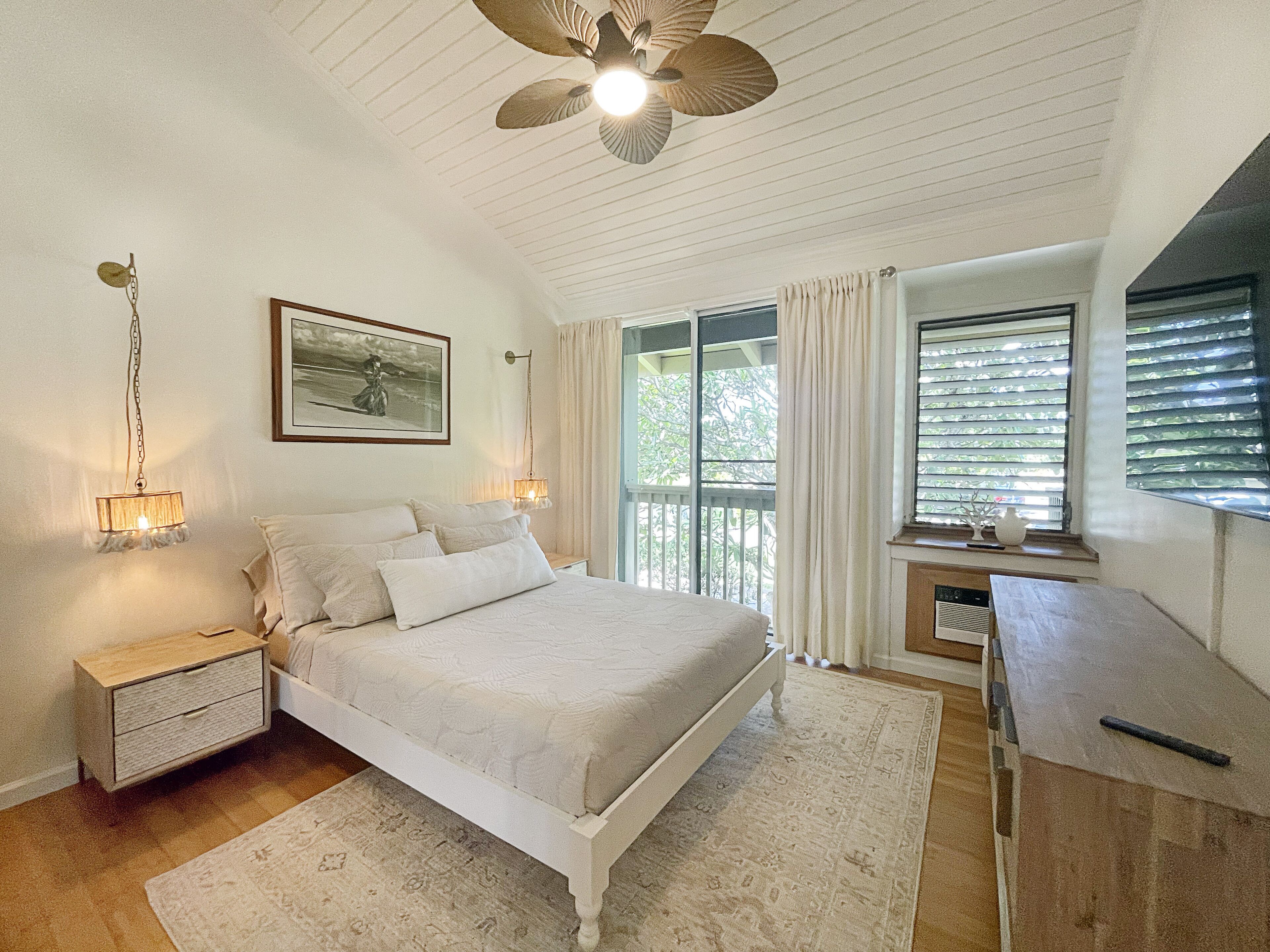 A bedroom with a bed, bedside lamps, a ceiling fan, a window with curtains, and a small table with a vase.