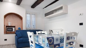 Un comedor con una mesa preparada para cuatro personas, sillas blancas, un sofá azul, un microondas y una pequeña zona de cocina con ventana.