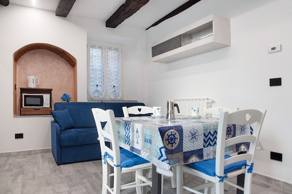 Un comedor con una mesa preparada para cuatro personas, sillas blancas, un sofá azul, un microondas y una pequeña zona de cocina con ventana.