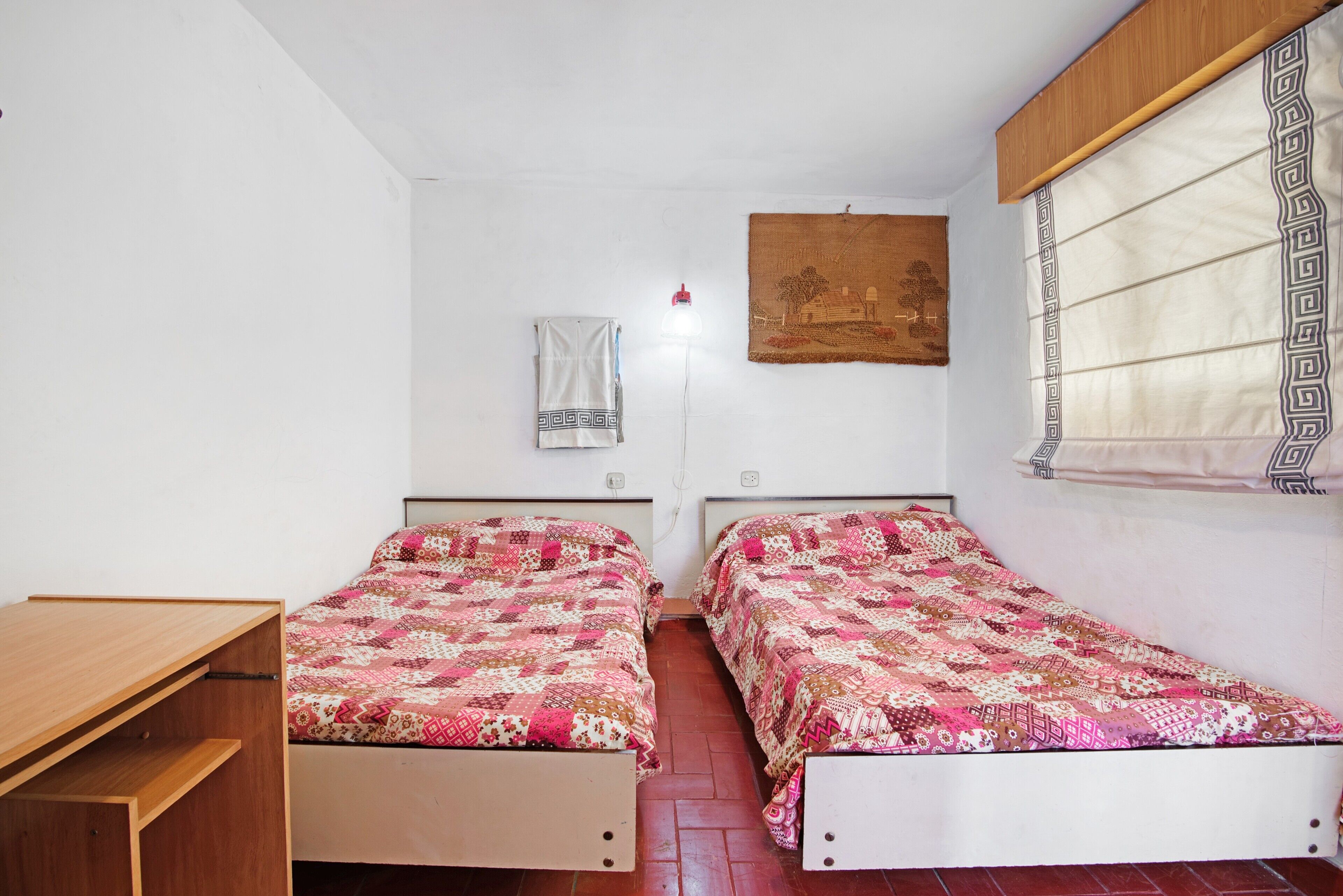 Une chambre avec deux lits, un bureau en bois, une lampe fixée au mur et une fenêtre ornée d’un rideau à motifs.