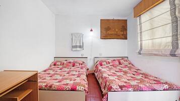 Une chambre avec deux lits, un bureau en bois, une lampe fixée au mur et une fenêtre ornée d’un rideau à motifs.