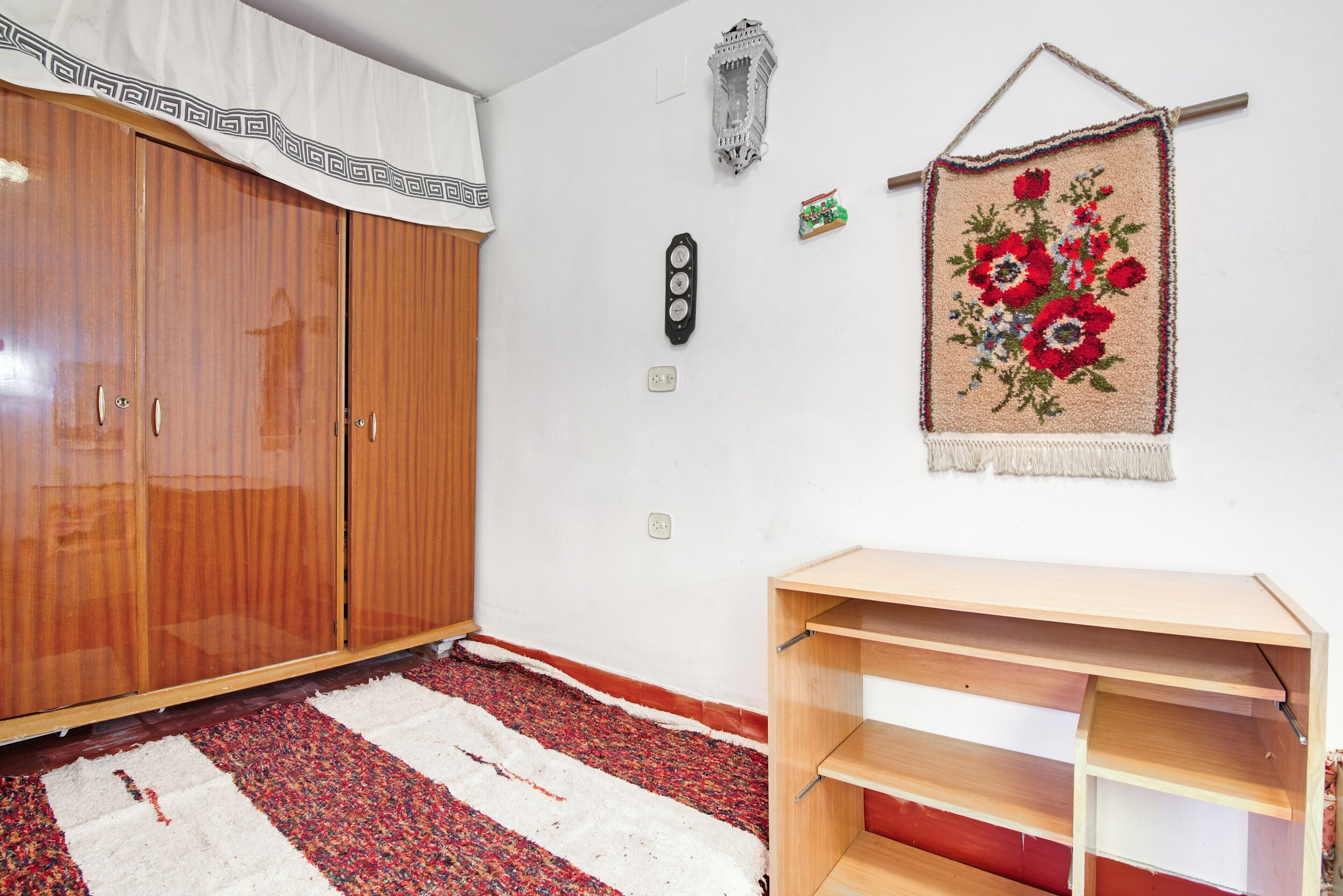 A room with a wooden wardrobe, a small wooden table, a patterned rug, a wall clock, and a tapestry.