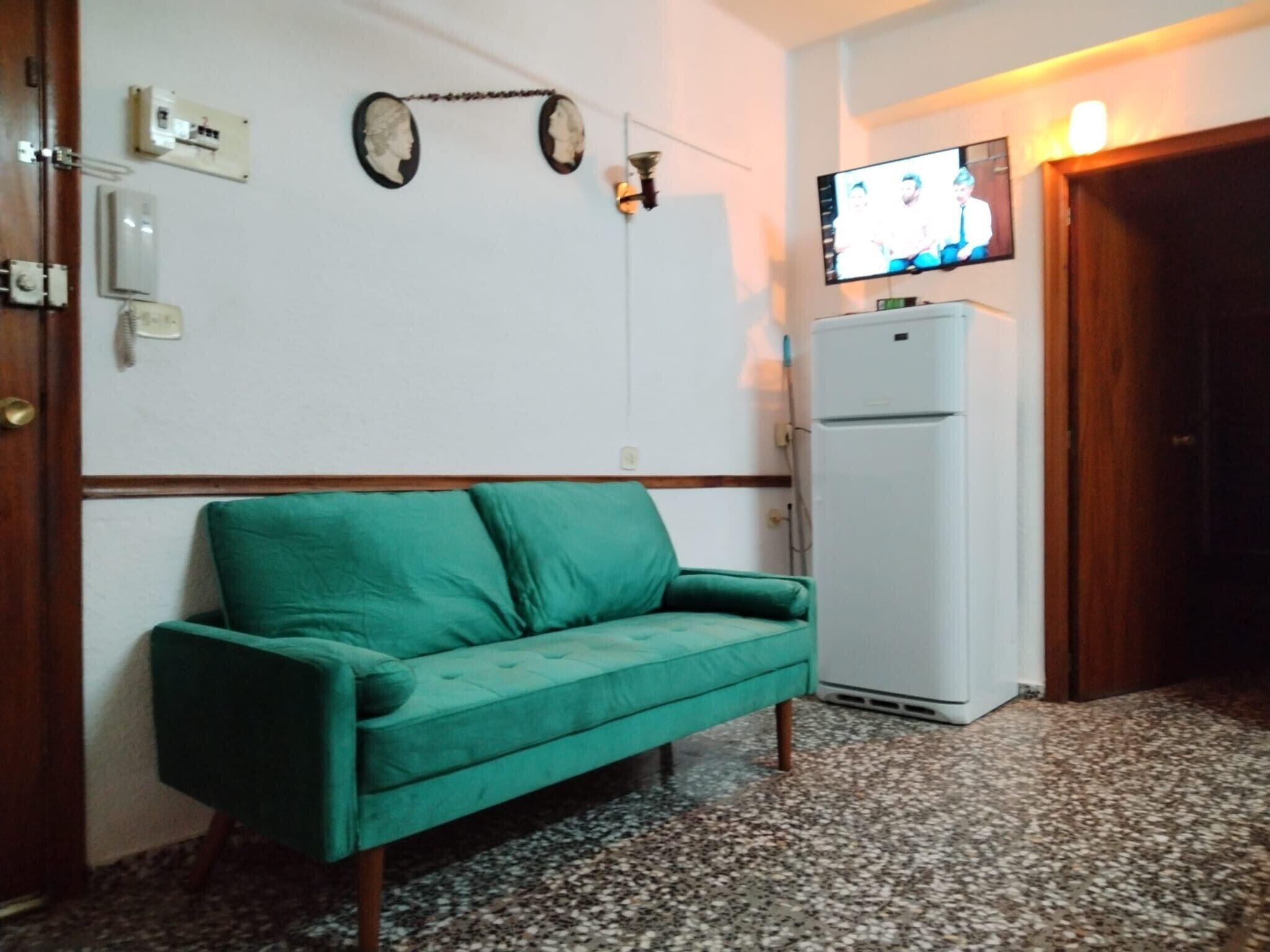 Habitació amb un sofà verd, un refrigerador blanc, una televisió penjada a la paret i una porta.