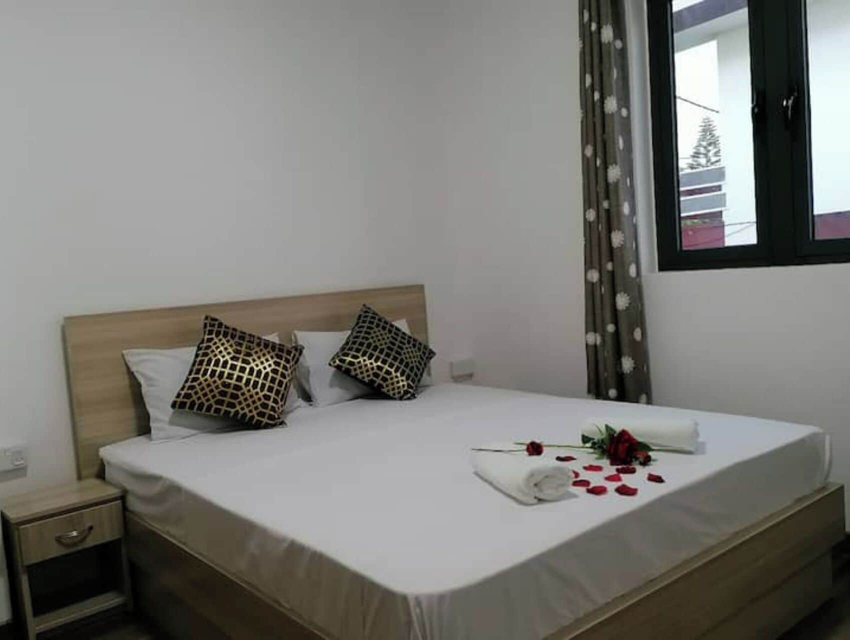 A neatly made bed with a white bedspread, two patterned pillows, a small bedside table with a drawer, and a window with a view of a building and trees.