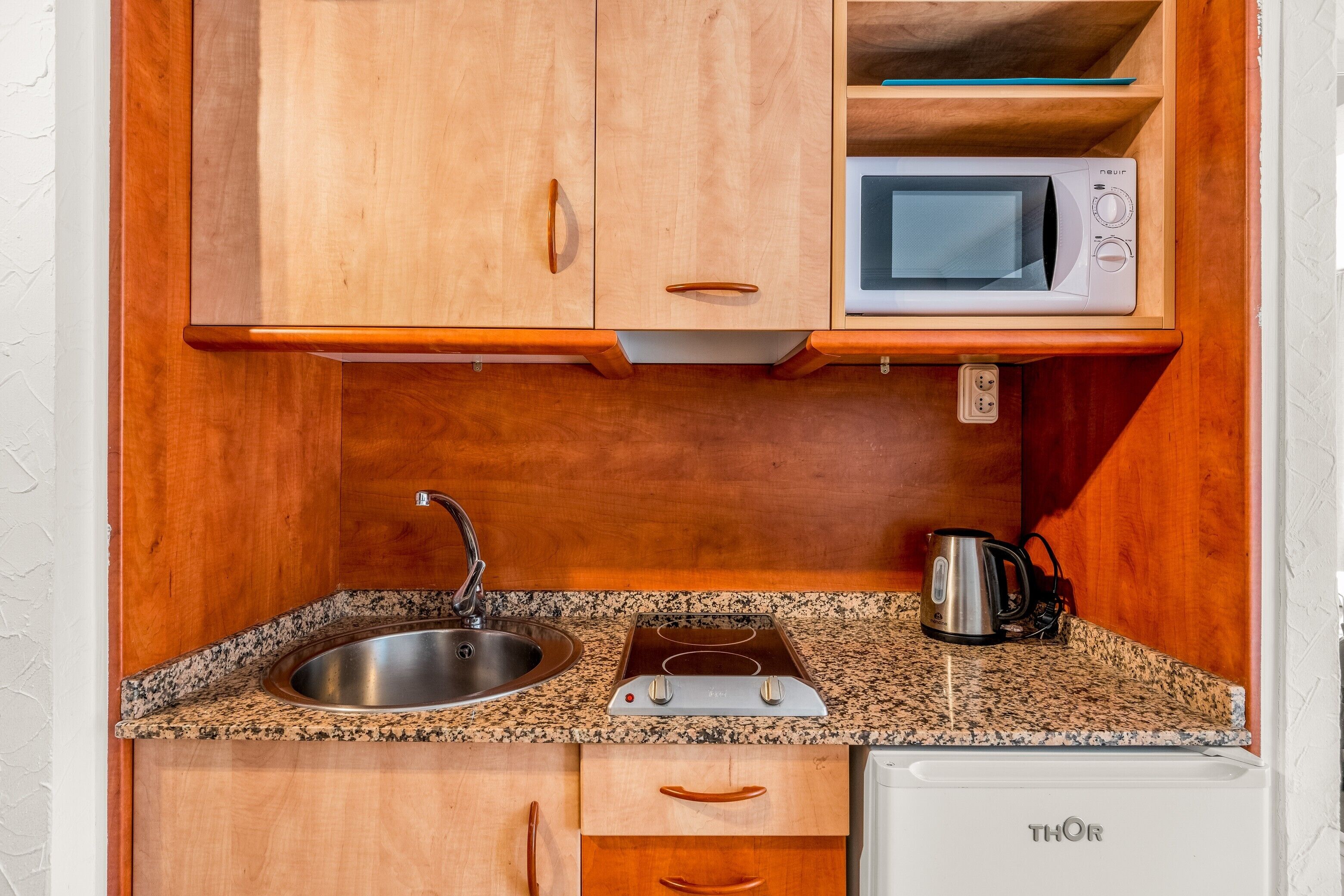 Una cocina compacta con armarios de madera, microondas integrado, pileta de acero inoxidable, hervidor y lavavajillas.