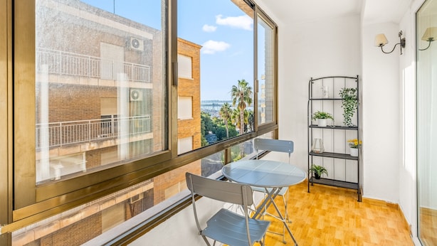 Un balcón con vistas a edificios y palmeras, una mesa y sillas de metal, y una estantería con plantas.