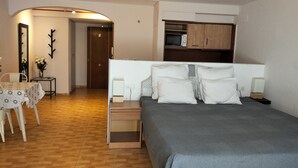Un dormitorio con cama, mesitas de noche, un escritorio con silla, un espejo y una puerta que da a otra habitación.