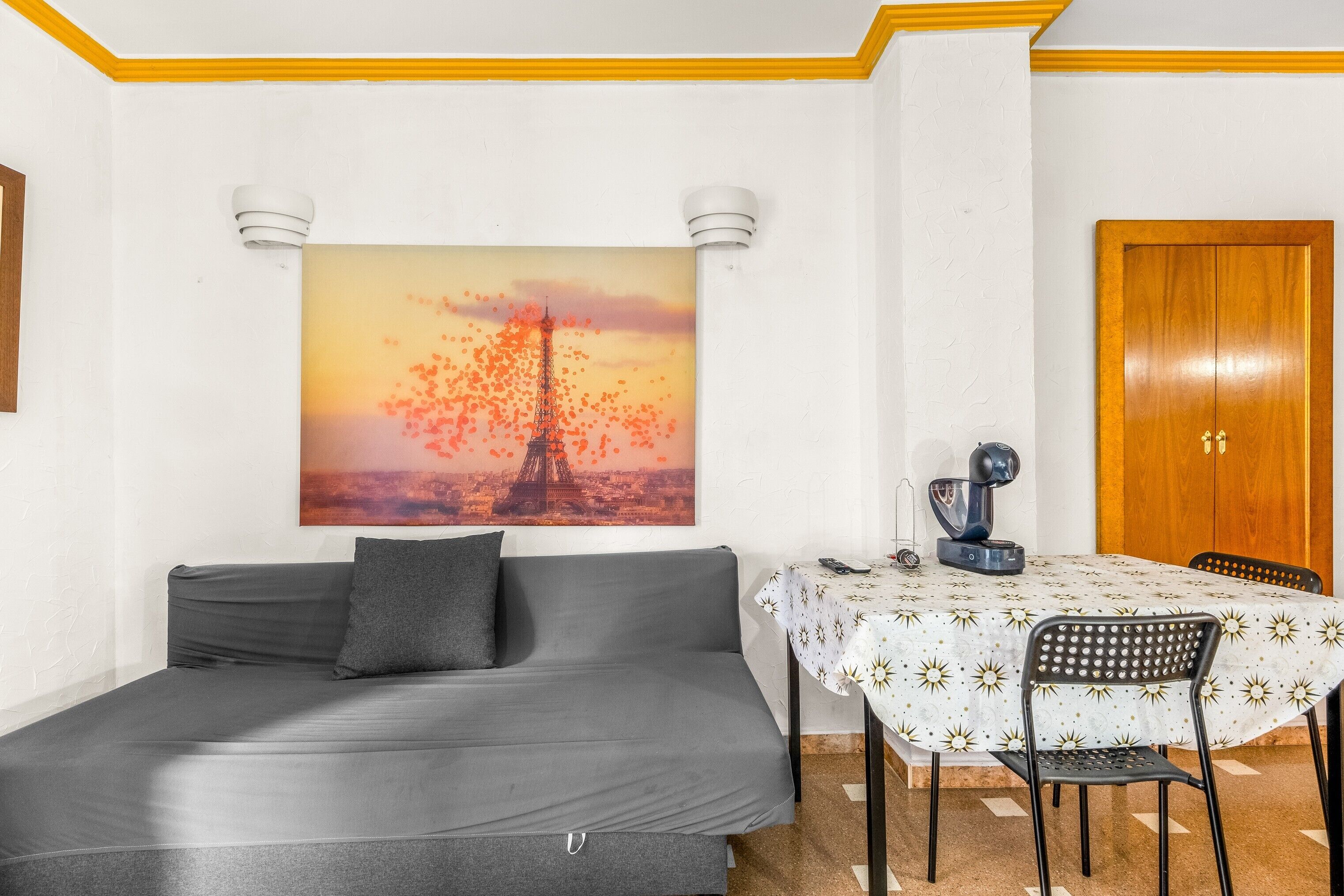 Una habitación con un sofá gris, una mesa de comedor con mantel blanco y un cuadro de la Torre Eiffel.
