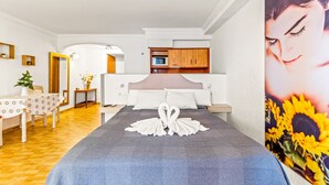 A hotel room with a bed, a dining table with a floral tablecloth, a small plant, and a kitchen area with wooden cabinets and a microwave.