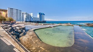 Un hotel frente al mar con piscina, costa rocosa y edificios al fondo.