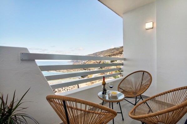 A balcony with wicker chairs, a table with a bottle and glass, and a view of the beach and hills.