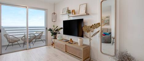A modern living room with a large TV, a sofa, a dining table, and a balcony with a view of the sea.