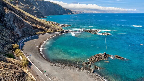 Un paisaje costero con una playa curva, afloramientos rocosos y una montaña al fondo.