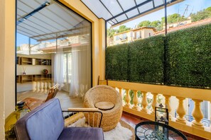 A balcony with a wicker chair, a glass door leading to a room with a sofa and a table, and a view of a neatly trimmed hedge and buildings.