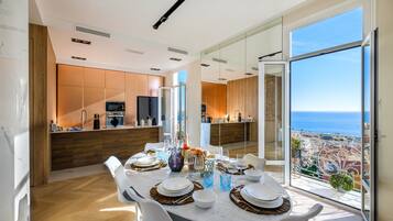 Una sala da pranzo moderna con un tavolo grande apparecchiato per sei persone, una cucina sullo sfondo e un balcone con vista sulla città e sul mare.